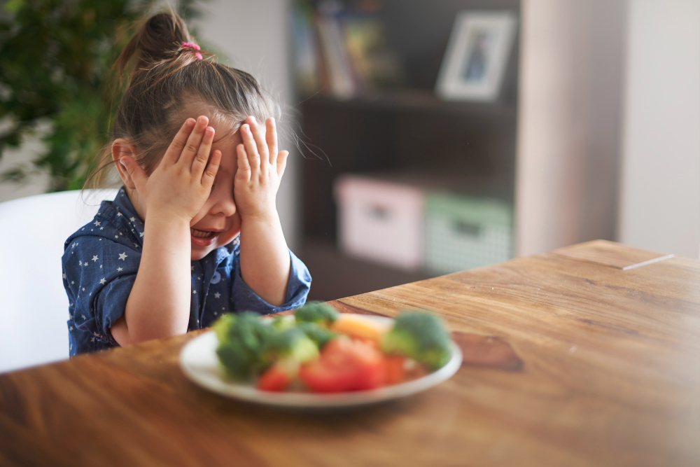 Young girl covering her eyes and refusing a plate of vegetables, a common picky eating behavior